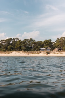 A serene coastal scene with gentle waves on the water in the foreground and a sandy beach lined with trees in the background. A few houses are nestled among the trees, offering a tranquil and secluded atmosphere. The sky above is partly cloudy with hints of blue.