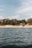 A serene coastal scene with gentle waves on the water in the foreground and a sandy beach lined with trees in the background. A few houses are nestled among the trees, offering a tranquil and secluded atmosphere. The sky above is partly cloudy with hints of blue.