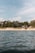 A serene coastal scene with gentle waves on the water in the foreground and a sandy beach lined with trees in the background. A few houses are nestled among the trees, offering a tranquil and secluded atmosphere. The sky above is partly cloudy with hints of blue.