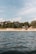 A serene coastal scene with gentle waves on the water in the foreground and a sandy beach lined with trees in the background. A few houses are nestled among the trees, offering a tranquil and secluded atmosphere. The sky above is partly cloudy with hints of blue.