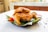 A plate of crispy, fried chicken is placed on a banana leaf. Accompanying the chicken are a tomato and cups of sauce. The dish is set on a white table with scattered black peppercorns adding decorative elements.