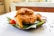 A plate of crispy, fried chicken is placed on a banana leaf. Accompanying the chicken are a tomato and cups of sauce. The dish is set on a white table with scattered black peppercorns adding decorative elements.