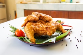A plate of crispy, fried chicken is placed on a banana leaf. Accompanying the chicken are a tomato and cups of sauce. The dish is set on a white table with scattered black peppercorns adding decorative elements.