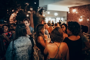 A group of people are gathered in an outdoor patio area, engaging in conversation and laughter. The setting has a warm and inviting atmosphere with string lights adding to the ambiance. Several individuals are facing each other, some wearing casual clothing like denim jackets and dresses. The background suggests an evening social event.