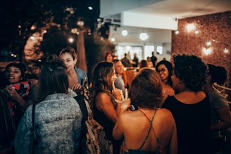 A group of people are gathered in an outdoor patio area, engaging in conversation and laughter. The setting has a warm and inviting atmosphere with string lights adding to the ambiance. Several individuals are facing each other, some wearing casual clothing like denim jackets and dresses. The background suggests an evening social event.