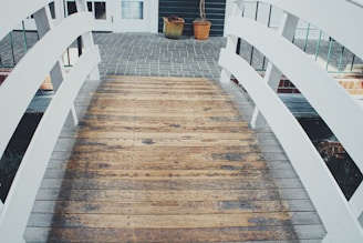 A wooden bridge with white railings leads to a paved area near a building entrance. Two potted plants are placed near the entrance, and there are glass windows and a door visible in the background.