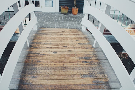 A wooden bridge with white railings leads to a paved area near a building entrance. Two potted plants are placed near the entrance, and there are glass windows and a door visible in the background.
