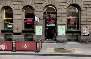 A brick facade with arched windows displaying neon signs for 'Bánh Mì' and 'Phở' adorns the exterior of a Vietnamese eatery. The entrance features a large menu and two large signs promoting catering services. Outdoor seating is partially visible, and there are promotional posters on the windows.