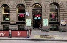 A brick facade with arched windows displaying neon signs for 'Bánh Mì' and 'Phở' adorns the exterior of a Vietnamese eatery. The entrance features a large menu and two large signs promoting catering services. Outdoor seating is partially visible, and there are promotional posters on the windows.