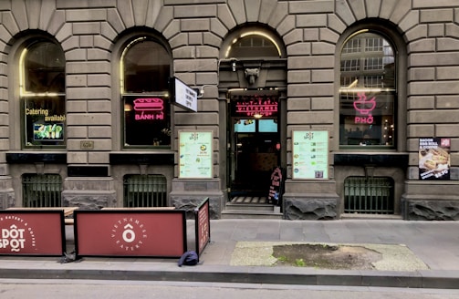 A brick facade with arched windows displaying neon signs for 'Bánh Mì' and 'Phở' adorns the exterior of a Vietnamese eatery. The entrance features a large menu and two large signs promoting catering services. Outdoor seating is partially visible, and there are promotional posters on the windows.