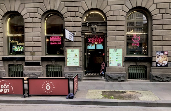 A brick facade with arched windows displaying neon signs for 'Bánh Mì' and 'Phở' adorns the exterior of a Vietnamese eatery. The entrance features a large menu and two large signs promoting catering services. Outdoor seating is partially visible, and there are promotional posters on the windows.