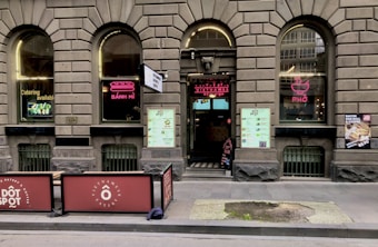 A brick facade with arched windows displaying neon signs for 'B&aacute;nh M&igrave;' and 'Phở' adorns the exterior of a Vietnamese eatery. The entrance features a large menu and two large signs promoting catering services. Outdoor seating is partially visible, and there are promotional posters on the windows.