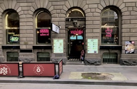 A brick facade with arched windows displaying neon signs for 'Bánh Mì' and 'Phở' adorns the exterior of a Vietnamese eatery. The entrance features a large menu and two large signs promoting catering services. Outdoor seating is partially visible, and there are promotional posters on the windows.