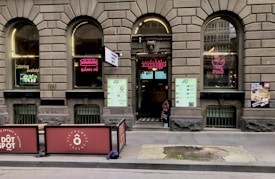 A brick facade with arched windows displaying neon signs for 'Bánh Mì' and 'Phở' adorns the exterior of a Vietnamese eatery. The entrance features a large menu and two large signs promoting catering services. Outdoor seating is partially visible, and there are promotional posters on the windows.