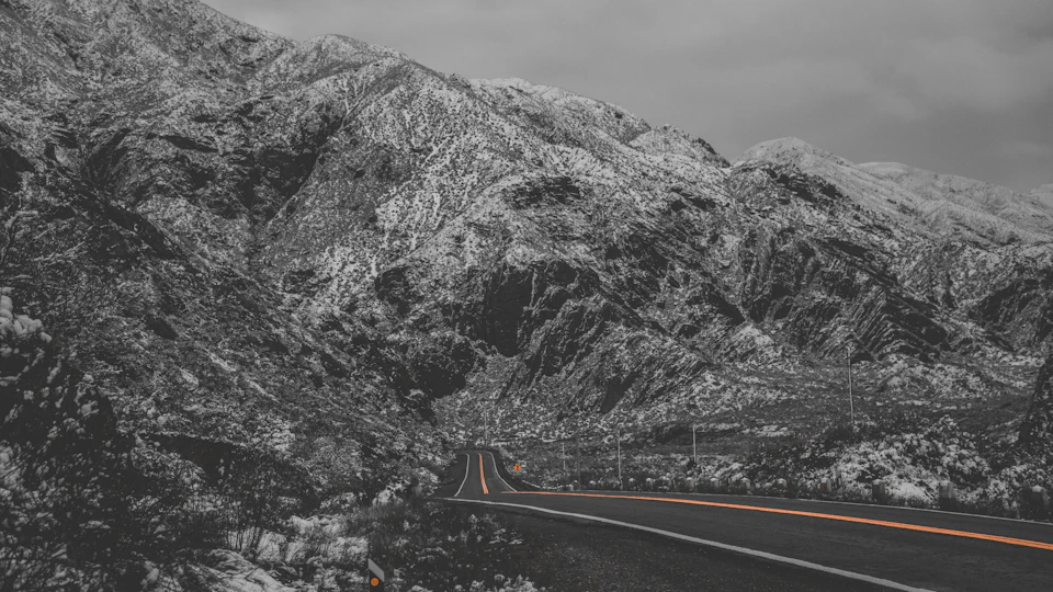 The image depicts a winding road cutting through a mountainous landscape, with snow-capped mountains rising up on either side. The sky above is overcast, casting a soft light over the scene and creating an ethereal atmosphere. The road curves gently to the right in the foreground, disappearing into the distance as it winds its way towards the horizon.

The mountains are blanketed in snow, their peaks dusted with white powder that glistens under the muted sunlight. The road is a stark contrast against this snowy backdrop, its black asphalt standing out prominently. In the background, there's an array of trees and shrubs, adding depth to the scene and providing a sense of scale as they recede into the distance.