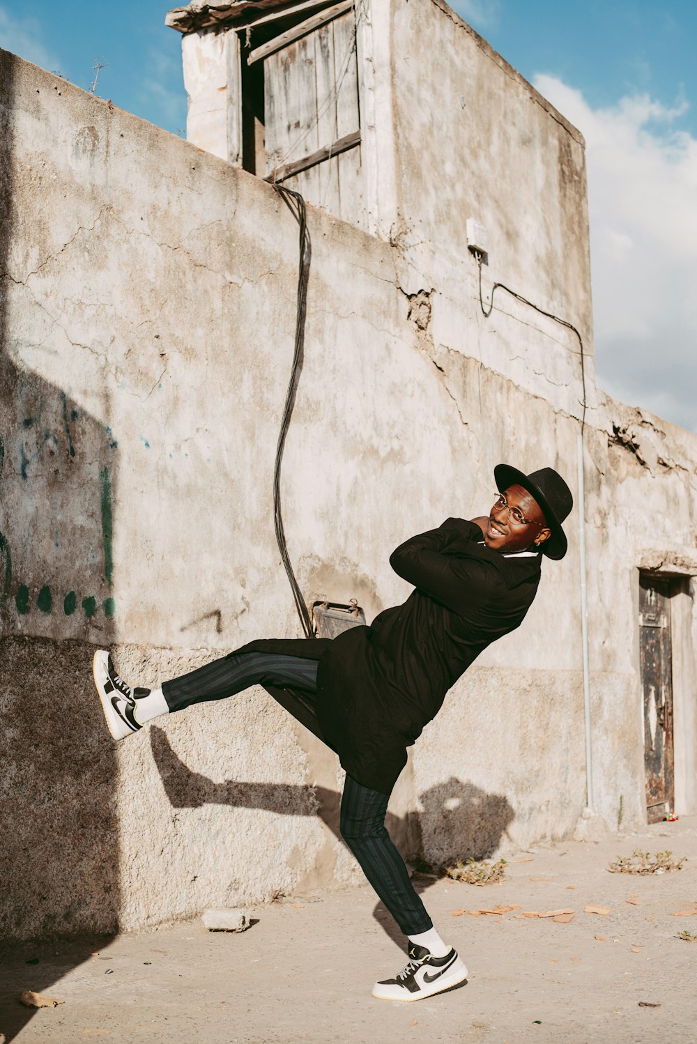 man in black jacket and black pants wearing black hat leaning on white wall during daytime