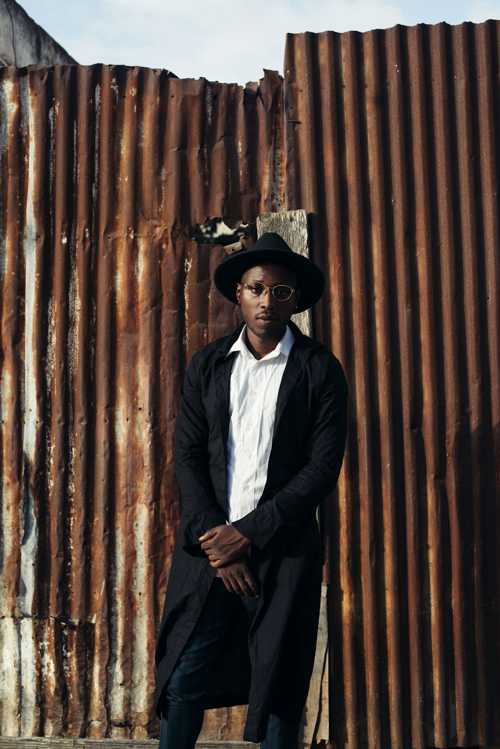 man in black suit jacket and black pants wearing black hat standing beside brown wooden wall