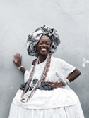 A woman dressed in elaborate traditional attire poses against a light gray wall. She wears a large, ornate headdress and a white, patterned dress adorned with beads and jewelry. Her smile is radiant, exuding confidence and joy. The attire is richly decorated, indicating cultural significance and celebration.