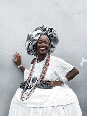 A woman dressed in elaborate traditional attire poses against a light gray wall. She wears a large, ornate headdress and a white, patterned dress adorned with beads and jewelry. Her smile is radiant, exuding confidence and joy. The attire is richly decorated, indicating cultural significance and celebration.