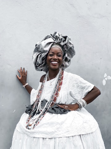 A woman dressed in elaborate traditional attire poses against a light gray wall. She wears a large, ornate headdress and a white, patterned dress adorned with beads and jewelry. Her smile is radiant, exuding confidence and joy. The attire is richly decorated, indicating cultural significance and celebration.