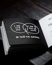 A book opened to a page featuring a black and white Venn diagram. The diagram is drawn with two overlapping circles containing Portuguese text. One circle says 'O que você ama' and the other 'O que te ama de volta'. The overlapping section, highlighted with an arrow pointing to it, forms part of the sentence 'Se você for sortudo.' on the page.