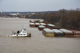 A towboat is navigating a river, pushing a long line of barges that are covered and arranged in rows. The river is calm, flanked by bare trees on one side and an industrial area with cranes and buildings on the other.