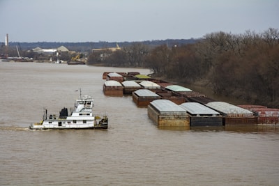 A towboat is navigating a river, pushing a long line of barges that are covered and arranged in rows. The river is calm, flanked by bare trees on one side and an industrial area with cranes and buildings on the other.