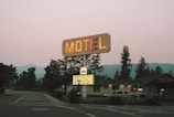 A vintage motel sign illuminated with neon lights stands alongside a road. Behind the sign, there is a one-story building that appears to be the motel. Tall trees and distant mountains are visible in the background as dusk approaches.