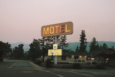 A vintage motel sign illuminated with neon lights stands alongside a road. Behind the sign, there is a one-story building that appears to be the motel. Tall trees and distant mountains are visible in the background as dusk approaches.