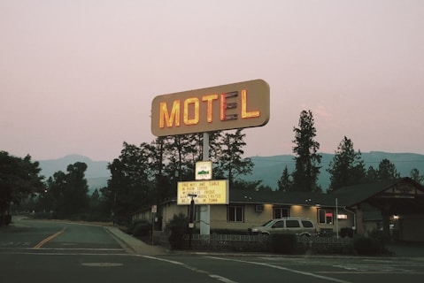 A vintage motel sign illuminated with neon lights stands alongside a road. Behind the sign, there is a one-story building that appears to be the motel. Tall trees and distant mountains are visible in the background as dusk approaches.
