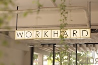 A rectangular sign with the words 'WORK HARD' is suspended from the ceiling. The background includes structural elements like pipes and beams, along with some greenery visible through windows.