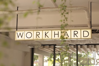 A rectangular sign with the words 'WORK HARD' is suspended from the ceiling. The background includes structural elements like pipes and beams, along with some greenery visible through windows.