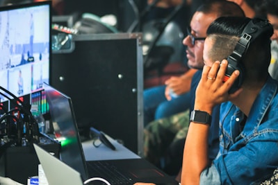 A person wearing headphones and a denim jacket is operating a control system at a media production station. Other individuals are visible in the background, possibly working together. There are multiple screens showing different video feeds and audio equipment visible on the table.