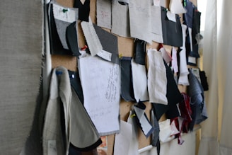 A corkboard covered with various fabric swatches pinned up. The swatches are of different colors, patterns, and textures, with some notes and writing visible on a piece of paper. The setup has a creative and organized appearance.