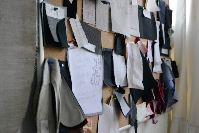 A corkboard covered with various fabric swatches pinned up. The swatches are of different colors, patterns, and textures, with some notes and writing visible on a piece of paper. The setup has a creative and organized appearance.