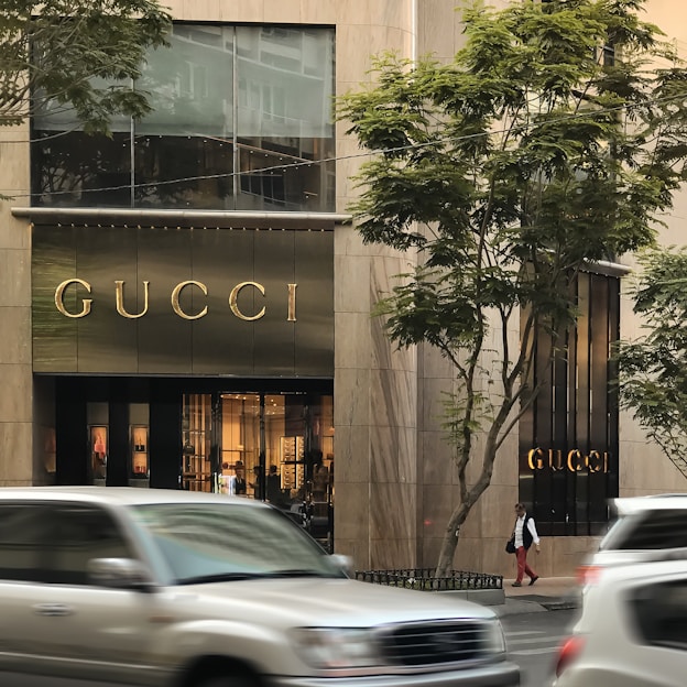 A storefront of a luxury brand with the name prominently displayed above the entrance. Large glass windows show elegant interior lighting and displays. Vehicles are blurred in motion on the street, and trees are visible on either side.