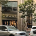 A storefront of a luxury brand with the name prominently displayed above the entrance. Large glass windows show elegant interior lighting and displays. Vehicles are blurred in motion on the street, and trees are visible on either side.