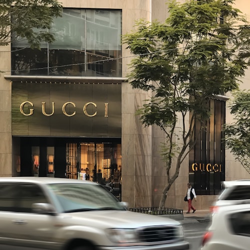 A storefront of a luxury brand with the name prominently displayed above the entrance. Large glass windows show elegant interior lighting and displays. Vehicles are blurred in motion on the street, and trees are visible on either side.