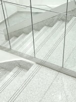 A staircase made of gray terrazzo and stainless steel lines, partially enclosed by transparent glass panels. The stair treads have several grooved lines running horizontally. The surrounding surfaces and the space appear clean and modern, with a minimalist design.