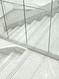 A staircase made of gray terrazzo and stainless steel lines, partially enclosed by transparent glass panels. The stair treads have several grooved lines running horizontally. The surrounding surfaces and the space appear clean and modern, with a minimalist design.