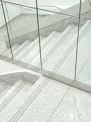 A staircase made of gray terrazzo and stainless steel lines, partially enclosed by transparent glass panels. The stair treads have several grooved lines running horizontally. The surrounding surfaces and the space appear clean and modern, with a minimalist design.