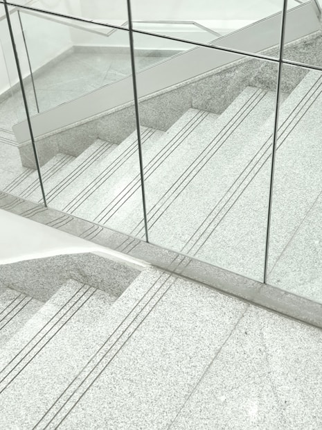 A staircase made of gray terrazzo and stainless steel lines, partially enclosed by transparent glass panels. The stair treads have several grooved lines running horizontally. The surrounding surfaces and the space appear clean and modern, with a minimalist design.