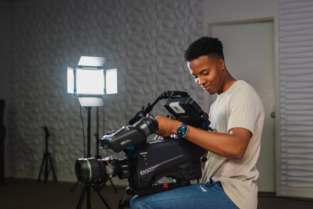 man in white t-shirt and blue denim jeans sitting on chair holding black dslr camera