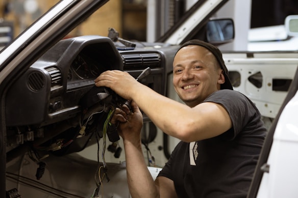 A person wearing a black t-shirt is smiling while working on the wiring of a car dashboard. The car's interior appears unfinished, showing exposed wires and missing panels.