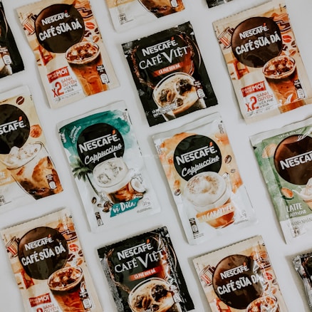 A collection of various Nescafé instant coffee packets arranged on a surface. The packets display different flavors and styles, including Café Sữa Đá, Café Việt, and Cappuccino, with colorful and distinct packaging designs.