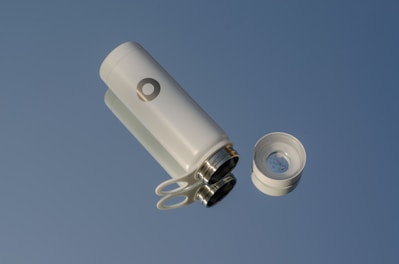 A modern, sleek white thermos with a cylindrical shape is positioned against a clear blue background. The thermos is shown with its cap off, revealing a stainless steel interior and an open top. The cap, which is also white with a metallic inside, lies next to the thermos.