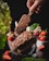 A hand holds a chocolate-covered ice cream bar with sprinkles above a glass bowl filled with other similar bars. Strawberries and leaves add a fresh, vibrant contrast to the rich tones of the chocolate.