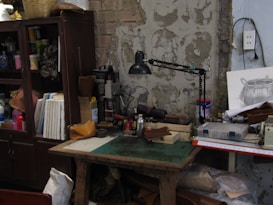 A cluttered workshop with a weathered wall in the background. The wooden desk is piled with tools and materials including a desk lamp, some papers, and small boxes. A drawing of a basket rests on the table. Nearby, a cabinet filled with various supplies and books adds to the crowded atmosphere. The environment feels practical and industrious, showing signs of being actively used.