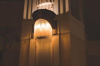 A decorative lamp with a vintage design, featuring multiple hanging beads or pendants, is mounted on a wall in an interior space. The warm, yellow light from the lamp casts soft shadows, creating an ambient and inviting atmosphere.