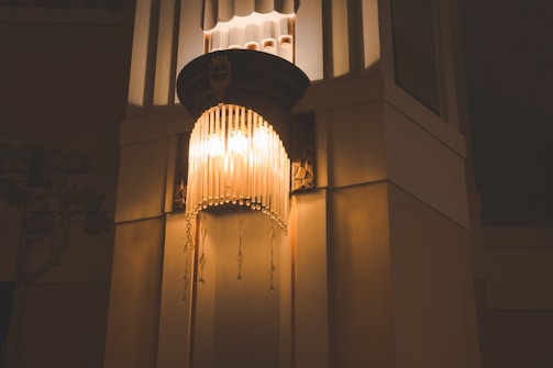 A decorative lamp with a vintage design, featuring multiple hanging beads or pendants, is mounted on a wall in an interior space. The warm, yellow light from the lamp casts soft shadows, creating an ambient and inviting atmosphere.